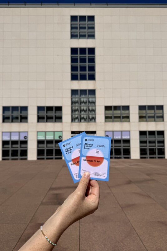 Hand hält zwei Tickets vor einem Gebäude mit vielen Fenstern