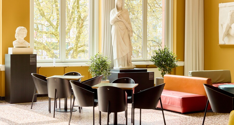 Café mit runden Tischen, schwarzen Stühlen, hohen Fenstern und weißen Skulpturen